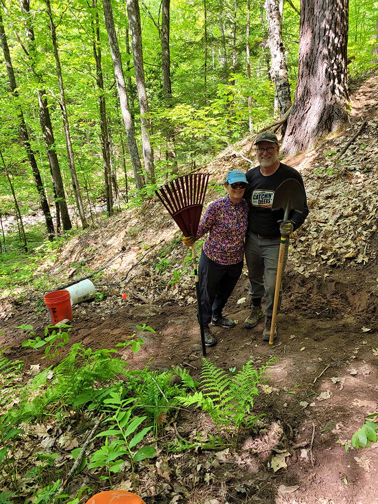 Two volunteers on a wooded hillside holding a rake and a shovel.